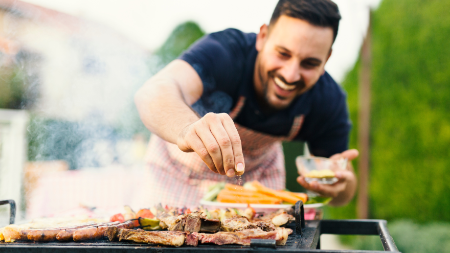 Lächelnder Mann würzt Fleisch am Grill im Garten.