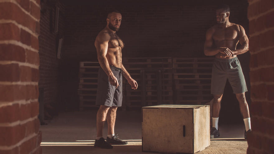 Two shirtless men in a rustic gym preparing for box jumps.