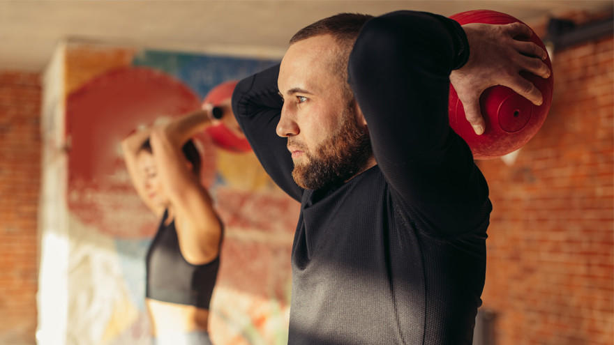 Focused man lifting medicine ball behind head during workout.