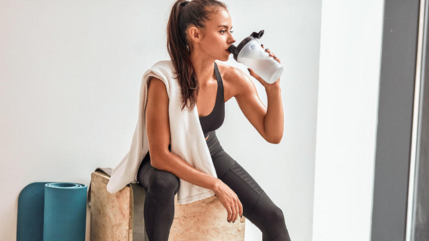 Vrouw drinkt eiwitshake na workout met fitnessmat op achtergrond.