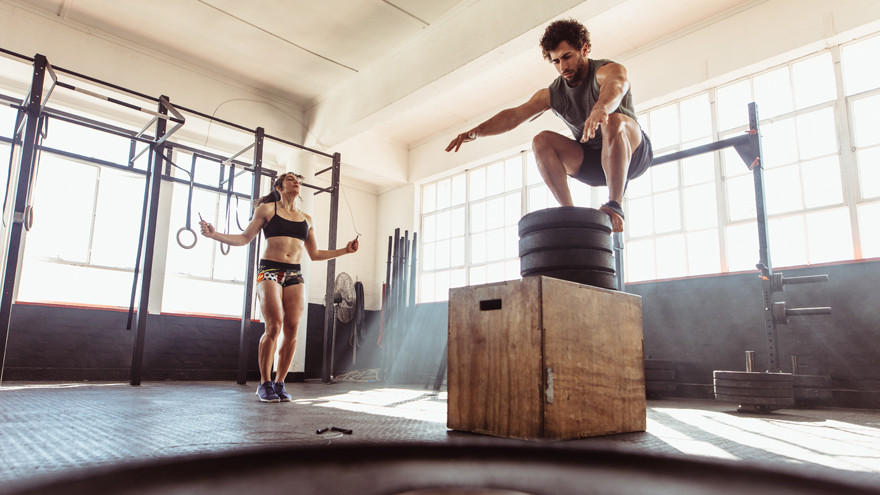 Man doet box jump, vrouw springt touw in gym.