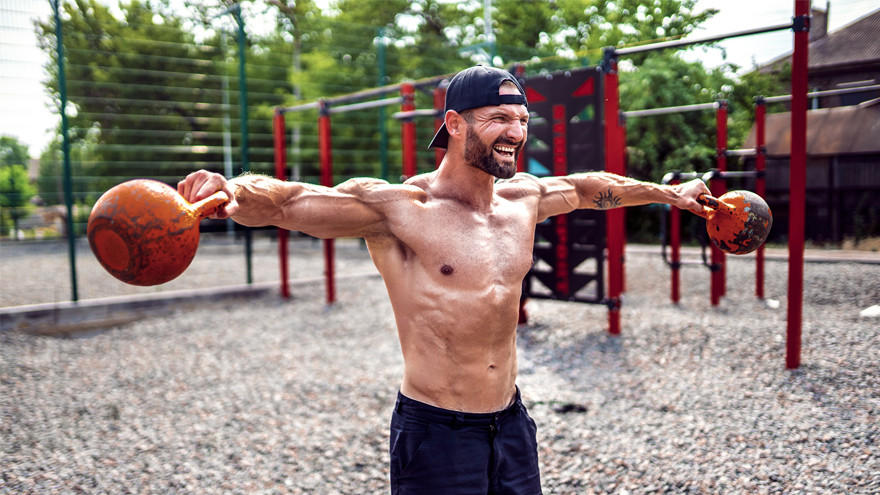 Homme torse nu soulevant des kettlebells en extérieur.