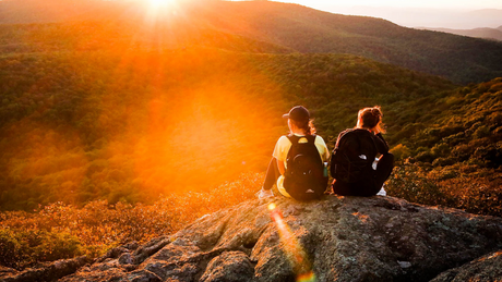 Two hikers resting on mountaintop with scenic forest view.