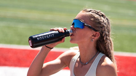 Sportlerin mit Sonnenbrille trinkt aus Body&Fit-Flasche.