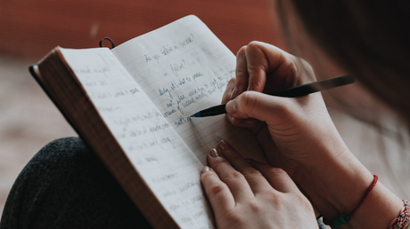 Frau schreibt mit Stift in ein aufgeschlagenes Notizbuch auf ihrem Schoß.