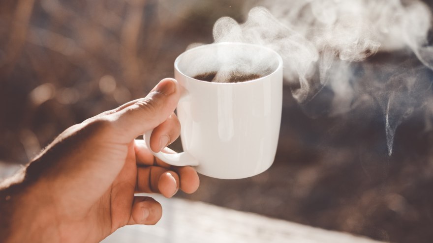 Hand hält dampfende Tasse Kaffee im Morgenlicht.