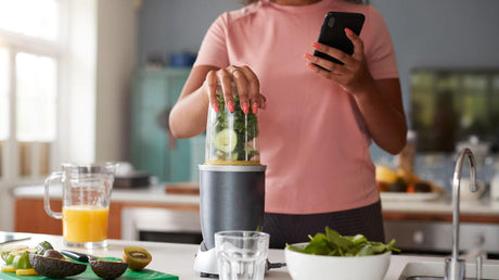Woman blending green smoothie while checking phone in kitchen.