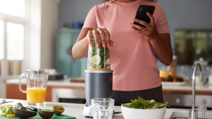 Vrouw maakt groene smoothie met blender in keuken.