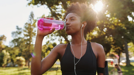 Femme qui boit de l'eau dans une gourde rose en extérieur après une course, avec des écouteurs et le soleil filtrant à travers les arbres