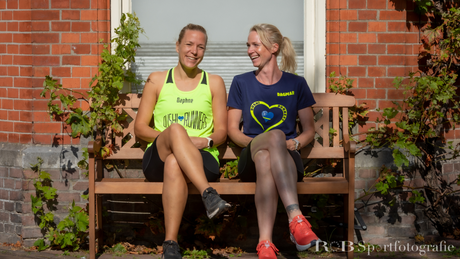 Twee vrouwen in sportkleding lachen op bankje buiten.