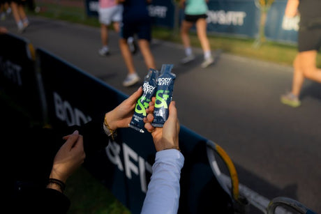 Hands holding two Body&Fit Energy Gel sachets at a race, runners passing on the course behind
