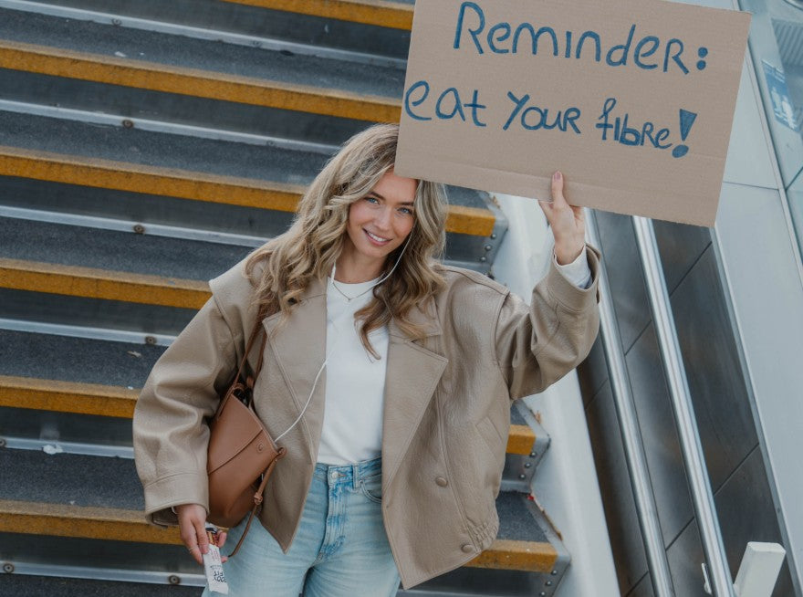 Woman on an escalator staircase holding a cardboard sign that reads 'Reminder: eat your fibre!'