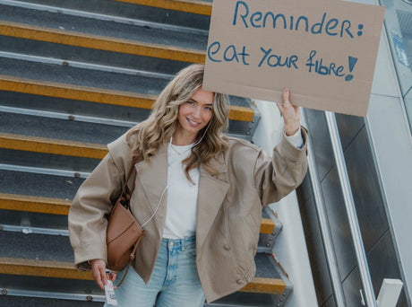 Woman on an escalator staircase holding a cardboard sign that reads 'Reminder: eat your fibre!'