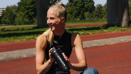 Smiling woman holding Body&Fit shaker on running track.