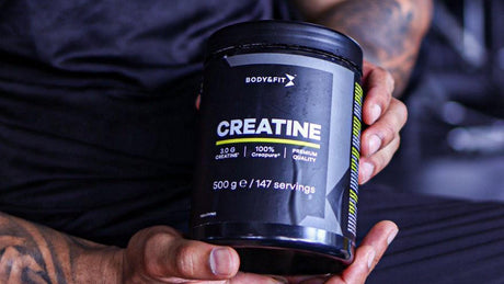 Body&Fit Kreatin-Dose wird in muskulösen Händen im Fitnessstudio präsentiert.