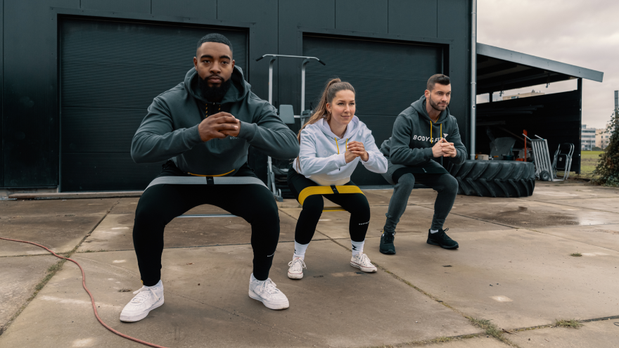Drie sporters doen squats met weerstandsband buiten.