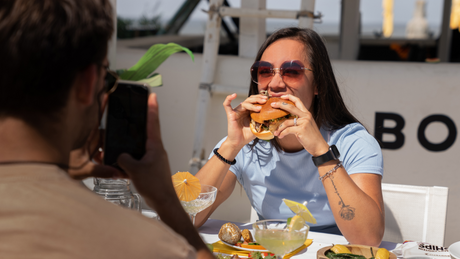 Frau genießt Burger auf Sommerterrasse, wird fotografiert.