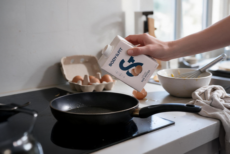 Une main verse Body&Fit Liquid Egg Whites de la bouteille dans une poêle chaude, avec une boîte d'œufs et un bol fouetté en arrière-plan.