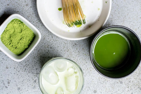 Matchapulver, Bambusbesen und eine Schale aufgeschlagener Matcha Tee neben einem Glas Milch mit Eis für einen Iced Matcha Latte