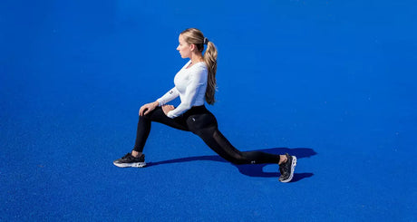 Woman doing deep lunge stretch on bright blue outdoor surface.
