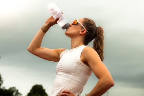 Femme en tenue de sport blanche buvant de l'eau à une gourde après l'entraînement, portant des lunettes de soleil sous un ciel nuageux.