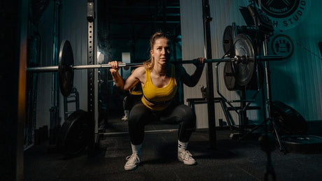 Femme en squat avec barre de musculation en salle.