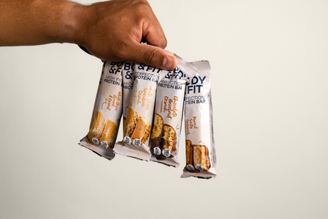 	Hand holding four Body&Fit protein bars, including Perfection Protein Bar in Chocolate Hazelnut Crunch, Peanut Salted Caramel and White Chocolate & Speculoos flavours