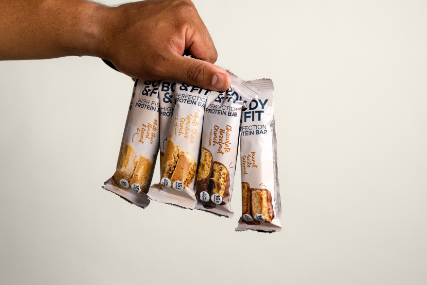 Hand hält vier Body&Fit Proteinriegel, darunter der Perfection Protein Bar in den Geschmacksrichtungen Chocolate Hazelnut Crunch, Peanut Salted Caramel und White Chocolate & Speculoos