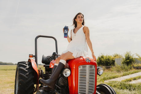 Frau im weißen Sommerkleid sitzt auf einem roten Traktor und hält einen Body&Fit Shaker auf einem Feld