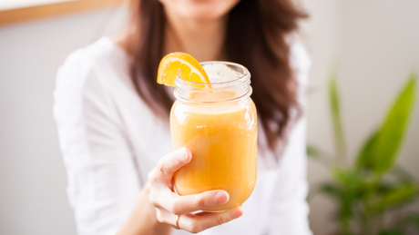 Vrouw houdt smoothie met sinaasappelstukje omhoog.