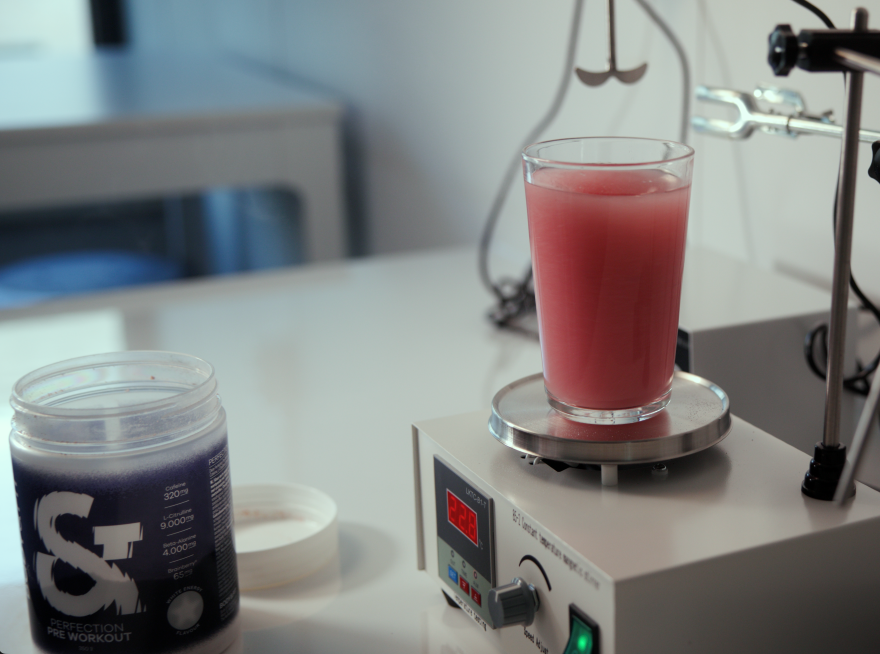 Glas Perfection Pre-Workout White Energy, durch Brainberry® rot gefärbt, auf einem Labor-Magnetrührer neben der geöffneten Dose