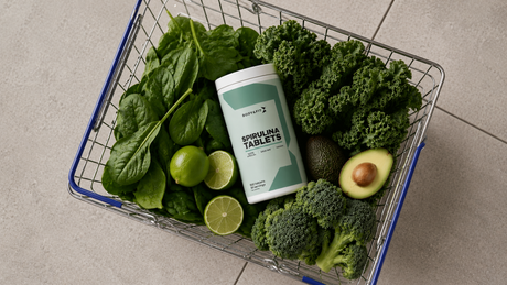 Panier de super-aliments verts comme les épinards, le chou kale, le brocoli, l'avocat et le citron vert, avec un pot de Spirulina Tablets de Body&Fit