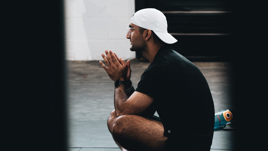 Man sitting cross-legged in gym, wearing cap and glasses.