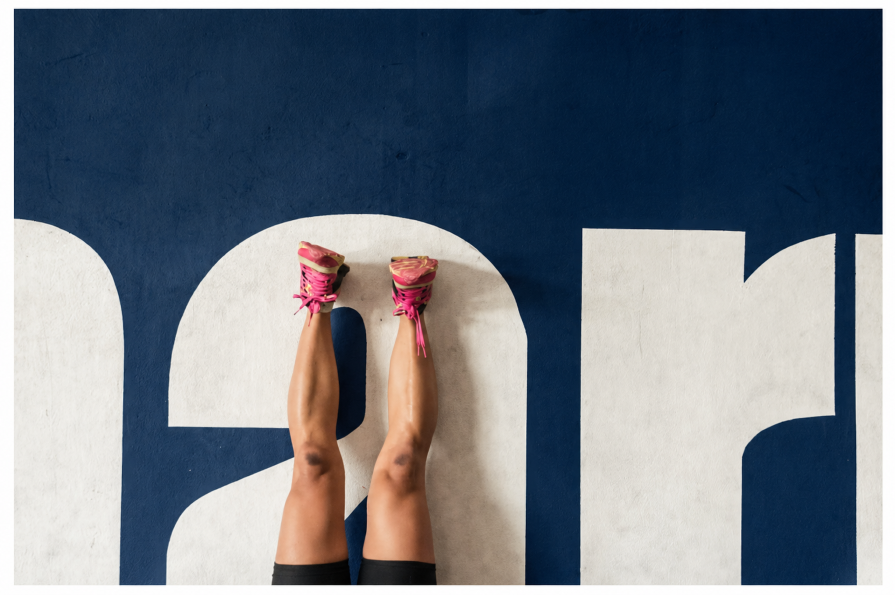 Top-down view of a person’s legs wearing pink sneakers, raised vertically against a dark blue wall with large white painted letters