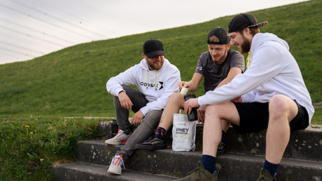 Drei junge Männer in sportlicher Freizeitkleidung sitzen gemeinsam auf Stufen mit Snacktüte.