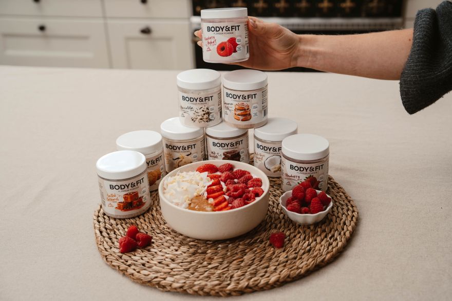 Yogurt bowl with raspberries, strawberries, coconut and white chocolate surrounded by Body&Fit Flavour Powder jars