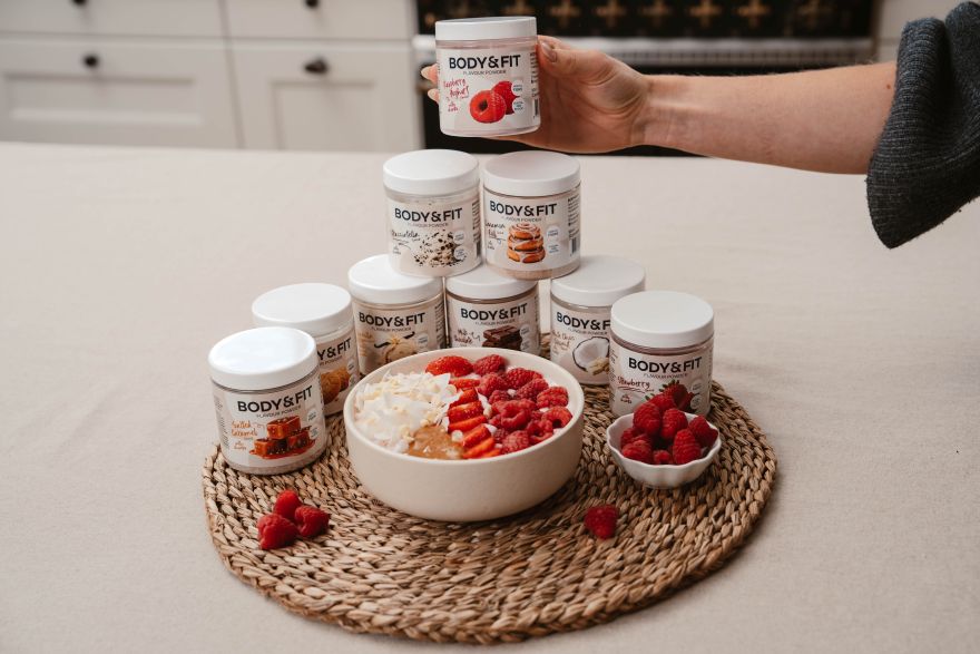 Yoghurt bowl met frambozen, aardbeien, kokos en witte chocolade tussen potten Body&Fit Flavour Powder