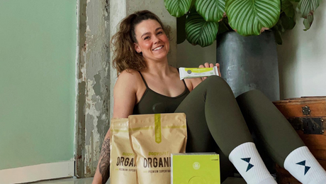Smiling woman sitting with Body&Fit organic supplements and protein bar.
