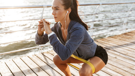 Woman doing resistance band squats on boardwalk by the sea.