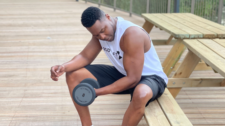 Man lifting dumbbell while seated on wooden bench outdoors.