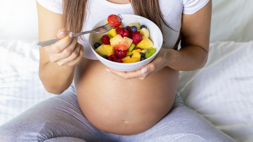 Zwangere vrouw eet kleurrijke fruitsalade uit witte kom.