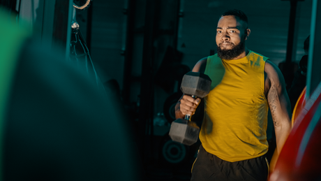Man lifting dumbbell in gym wearing yellow tank top.