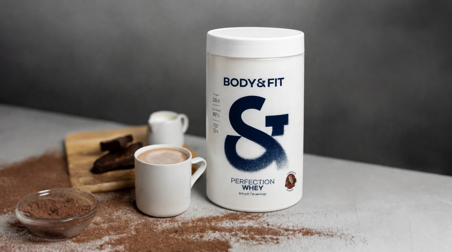 Protéine whey Body&Fit à côté d’une tasse de chocolat chaud avec du cacao en poudre et des morceaux de chocolat