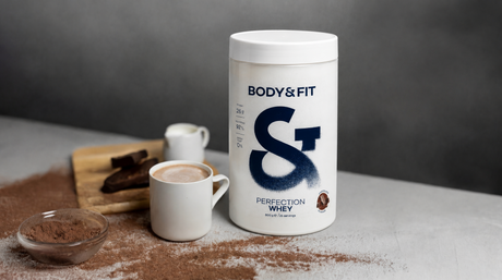 Protéine whey Body&Fit à côté d’une tasse de chocolat chaud avec du cacao en poudre et des morceaux de chocolat