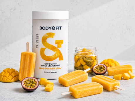 Pot de Body&Fit Perfection Whey Lemonade saveur Mangue-Fruit de la Passion, entouré de glaces protéinées, de mangue fraîche et de fruit de la passion.