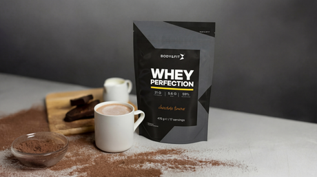 Body&Fit whey proteïne naast een kop warme chocolademelk met cacaopoeder en stukjes chocolade