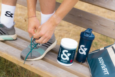 Man tying running shoe laces on a bench next to Body&Fit Perfection Pre-Workout tub, shaker, and gym bag