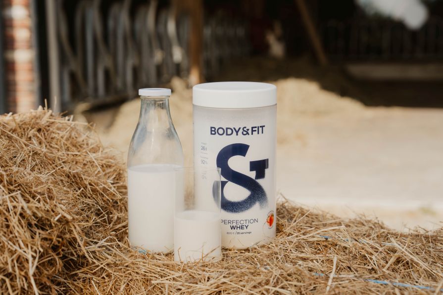Body&Fit Perfection Whey Proteindose mit einem Glas Milch und gemixtem Shake auf einem Heuballen, Bauernhof im Hintergrund.
