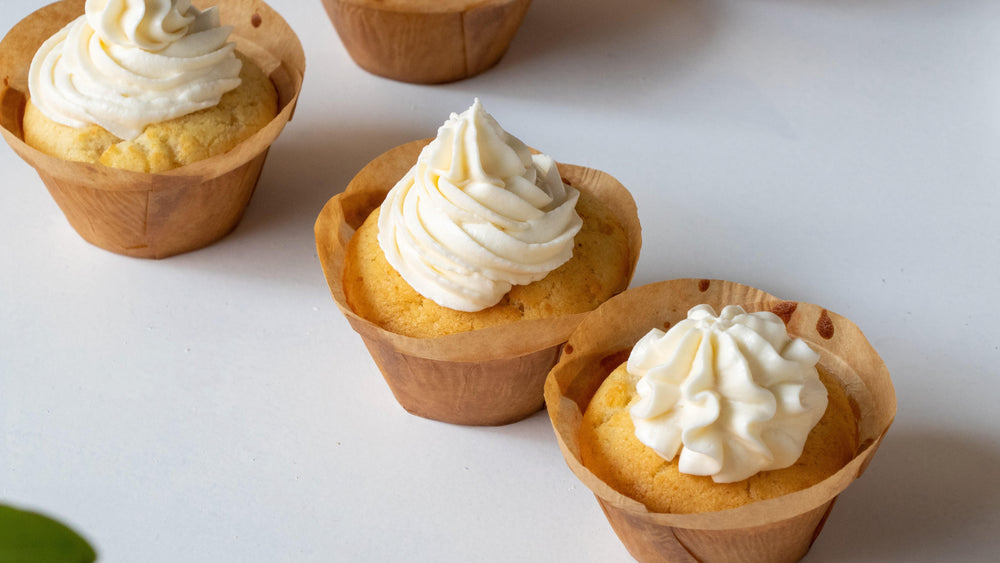 Kleine Vanille-Cupcakes mit Sahnehaube in braunen Papierförmchen auf weißem Hintergrund.