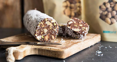 Schoko-Nuss-Salami auf Holzbrett angeschnitten, Body&Fit Verpackung im Hintergrund.